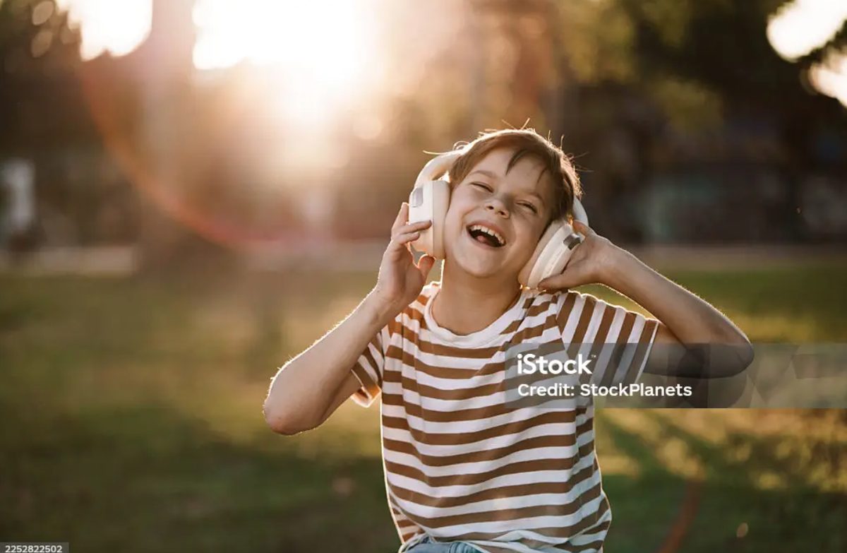 A boy laughing with delight, hands on his headphones, lit by golden evening sun in a park