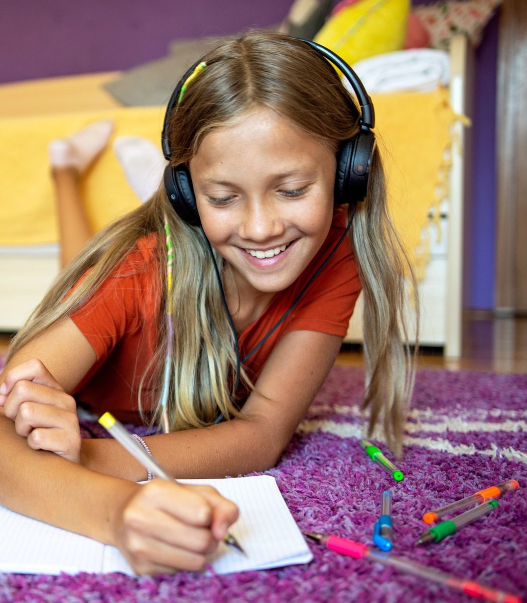 A girl wearing headphones drawing in a notebook on a purple rug